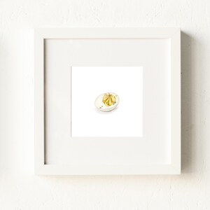 May include: A framed watercolor print of a deviled egg on a small white plate. The egg is cut in half, with a yellow yolk filling and a garnish of green herbs. The frame is white.