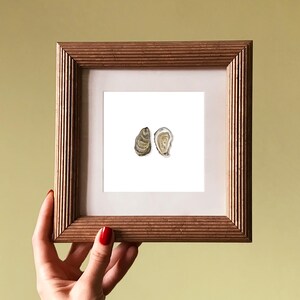 May include: A framed watercolor print featuring two oyster shells. The artwork is set against a white background, framed in a square, ribbed, wooden frame. The frame is held by a hand with red nail polish.