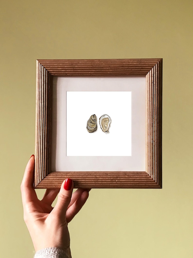 May include: A framed watercolor print featuring two oyster shells. The artwork is set against a white background, framed in a square, ribbed, wooden frame. The frame is held by a hand with red nail polish.