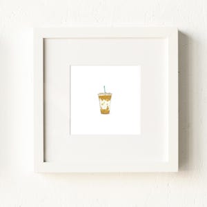 May include: A framed print featuring a watercolor illustration of an iced coffee. The coffee is in a clear cup with a blue straw. The drink has a light brown color with white accents. The frame is white and the background is white.
