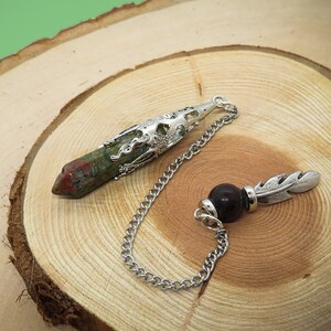 May include: Two crystal pendulums on a wooden surface. One pendulum is a green, red, and brown crystal with a silver top. The other pendulum is a dark purple crystal with a silver feather.