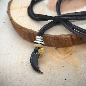 Può includere: Una collana in corda di cuoio marrone con un ciondolo a forma di artiglio di orso nero e una perla a righe.