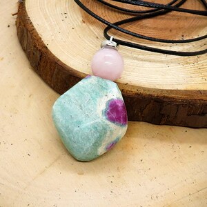 Pode incluir: Um pingente de pedra preciosa verde e rosa em um cordão de couro preto. O pingente tem uma forma irregular e bruta, com uma base verde claro e inclusões rosa e brancas. O cordão é amarrado no topo do pingente.