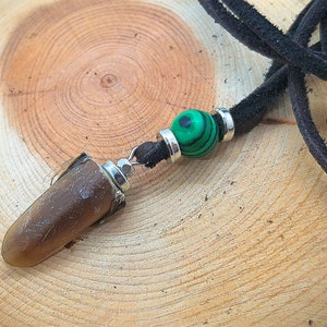 Puede incluir: Un colgante marrón en forma de lágrima cuelga de un cordón de cuero negro. El colgante está unido a un anillo plateado con una cuenta de malaquita verde.