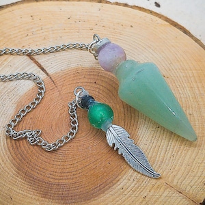 May include: Two pendulums with chains. One pendulum has a green and black crystal with a silver feather charm. The other pendulum has a light green crystal with a purple crystal at the top.