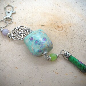 Puede incluir: Llavero artesanal con una piedra turquesa (aprox. 2,5 cm) con manchas rosas, un dije de nudo celta plateado y un colgante de cristal verde y morado. Incluye un cierre y cuentas plateadas.