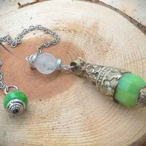 May include: A silver-toned pendulum with a green faceted bead and a decorative metal end. The pendulum has a silver chain and a clear, round bead. The decorative end has an ornate design and a green, faceted bead.