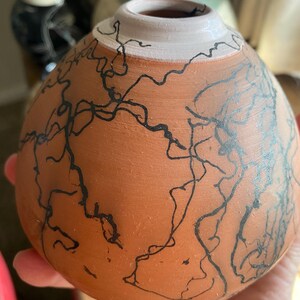 May include: A terracotta-colored ceramic vase with a white rim and black abstract line art. The vase has a rounded shape and is held in a hand. Other ceramic vases are visible in the background.