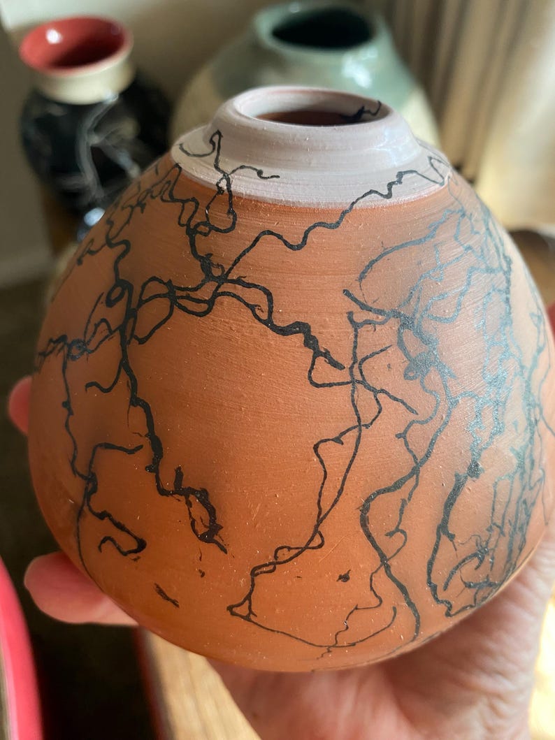 May include: A terracotta-colored ceramic vase with a white rim and black abstract line art. The vase has a rounded shape and is held in a hand. Other ceramic vases are visible in the background.