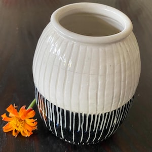 May include: A handcrafted ceramic vase with a white upper half and a black lower half. The white section has vertical textured lines, while the black section features vertical white lines. An orange flower rests beside the vase.