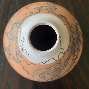 May include: A handmade ceramic vase with a round body and a wide opening. The vase is a warm terracotta color with a white interior rim. Dark blue, abstract lines decorate the exterior, creating a unique, artistic design.