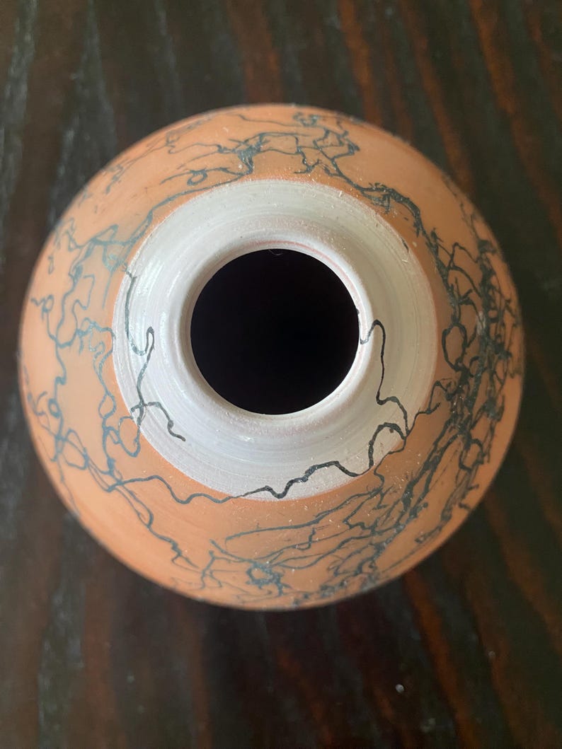 May include: A handmade ceramic vase with a round body and a wide opening. The vase is a warm terracotta color with a white interior rim. Dark blue, abstract lines decorate the exterior, creating a unique, artistic design.