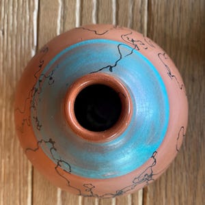 May include: A handmade terracotta-colored ceramic vase with a turquoise blue band and black line designs. The vase has a round shape with a small opening at the top. The interior is dark.