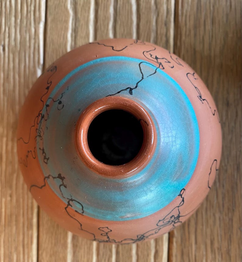 May include: A handmade terracotta-colored ceramic vase with a turquoise blue band and black line designs. The vase has a round shape with a small opening at the top. The interior is dark.