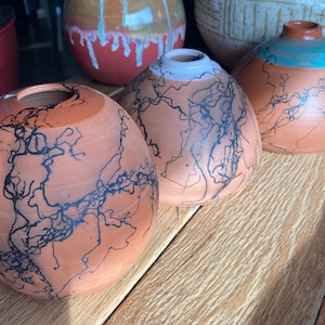 May include: Handmade terracotta vases with a unique lightning pattern. The vases are a warm orange color with black lines and a smooth finish. The vases are round with a small opening at the top. The vases are displayed on a wooden surface.
