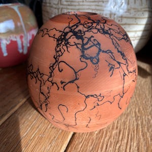 May include: A round, terracotta-colored ceramic vase with a black, lightning-like design. The vase has a small opening at the top and sits on a wooden surface. Other ceramic vases are visible in the background.