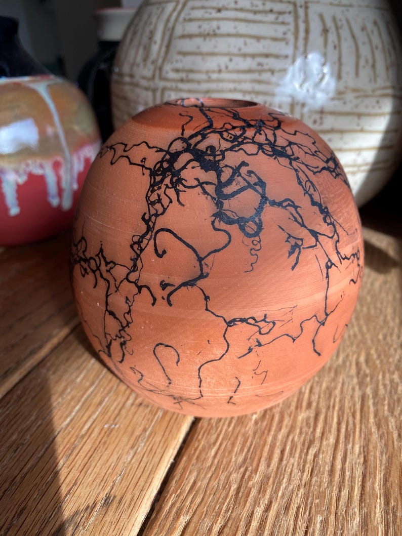 May include: A round, terracotta-colored ceramic vase with a black, lightning-like design. The vase has a small opening at the top and sits on a wooden surface. Other ceramic vases are visible in the background.