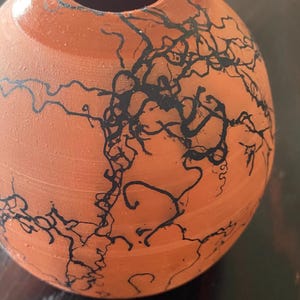 May include: A small, round, terracotta-colored ceramic vase. The vase is adorned with intricate black, lightning-like patterns across its surface. The design gives the vase a unique, artistic appearance. The vase has a smooth, unglazed finish.