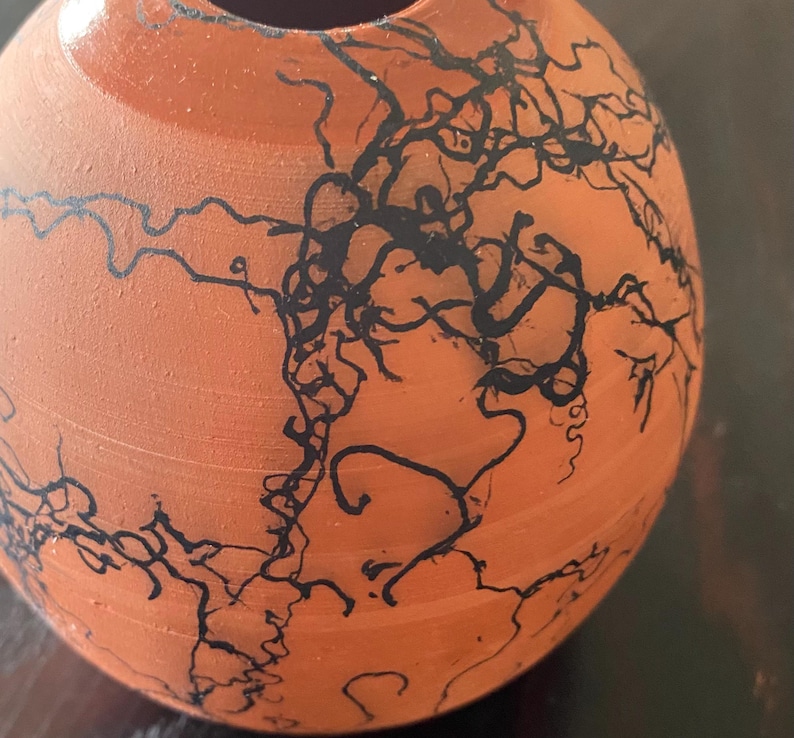 May include: A small, round, terracotta-colored ceramic vase. The vase is adorned with intricate black, lightning-like patterns across its surface. The design gives the vase a unique, artistic appearance. The vase has a smooth, unglazed finish.