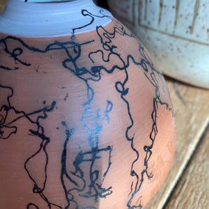 May include: A terracotta-colored ceramic vase with a light pink rim and black abstract line art. The vase has a rounded body and a narrow neck. Another ceramic piece is visible in the background.