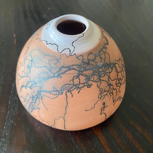 May include: A handmade ceramic vase with a unique design. The vase has a rounded shape with a white rim and a dark interior. The exterior is a warm orange color, decorated with black, lightning-like patterns.