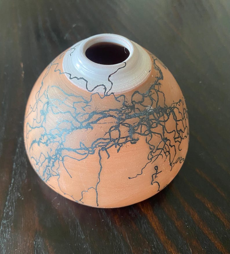 May include: A handmade ceramic vase with a unique design. The vase has a rounded shape with a white rim and a dark interior. The exterior is a warm orange color, decorated with black, lightning-like patterns.