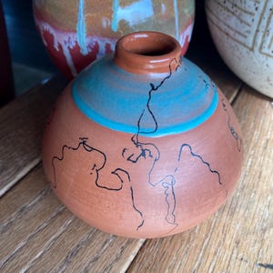 May include: A small, round terracotta vase with a narrow neck. The vase features a turquoise band and black line art. Other ceramic vases are visible in the background, suggesting a pottery collection.