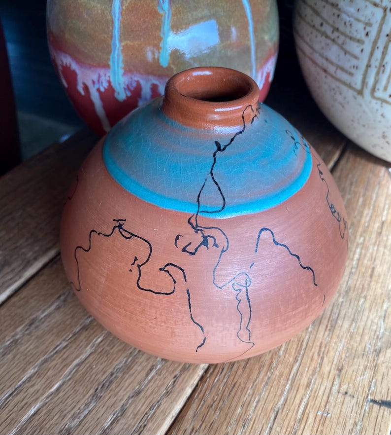 May include: A small, round terracotta vase with a narrow neck. The vase features a turquoise band and black line art. Other ceramic vases are visible in the background, suggesting a pottery collection.