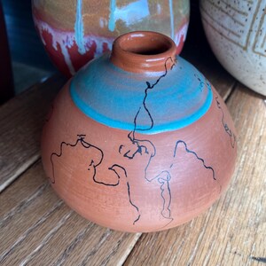 May include: A small, round terracotta vase with a narrow neck. The vase features a turquoise band and black line art. Other ceramic vases are visible in the background, suggesting a pottery collection.