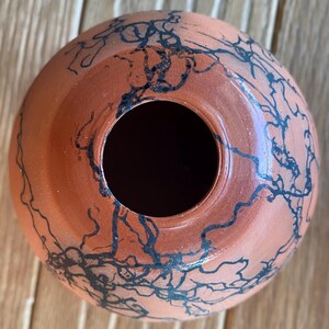 May include: A terracotta-colored ceramic vase with a round shape and a wide opening. The vase is decorated with black, abstract, linear designs. The vase sits on a wooden surface.