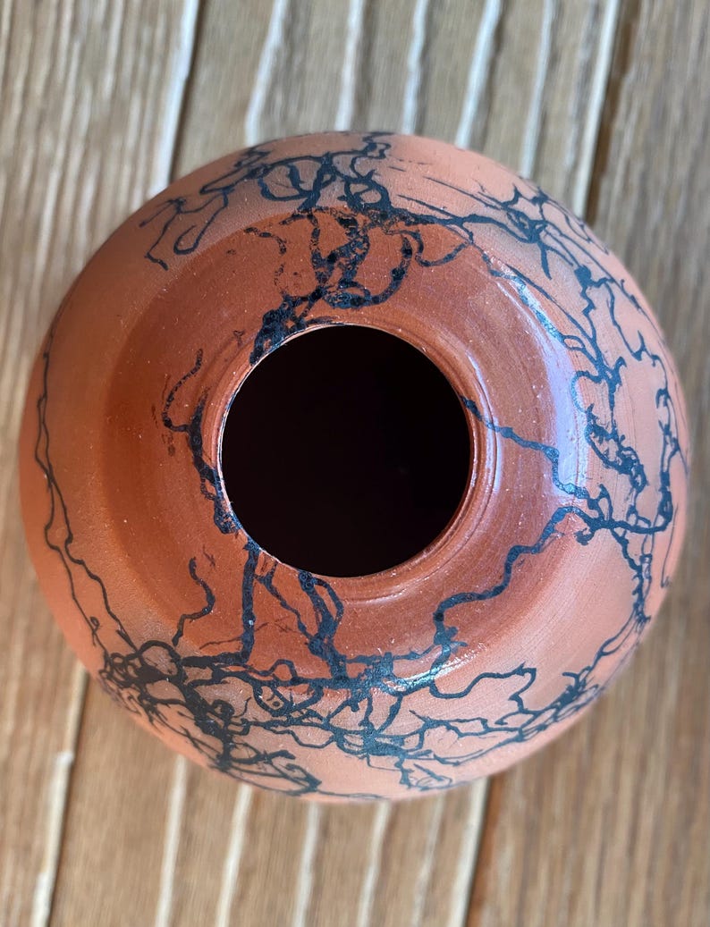 May include: A terracotta-colored ceramic vase with a round shape and a wide opening. The vase is decorated with black, abstract, linear designs. The vase sits on a wooden surface.