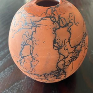 May include: A small, round terracotta vase with a narrow opening. The vase is decorated with a black, intricate, lightning-like pattern across its surface. The color is a warm orange-brown.