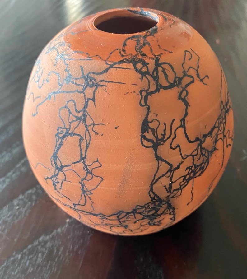 May include: A small, round terracotta vase with a narrow opening. The vase is decorated with a black, intricate, lightning-like pattern across its surface. The color is a warm orange-brown.