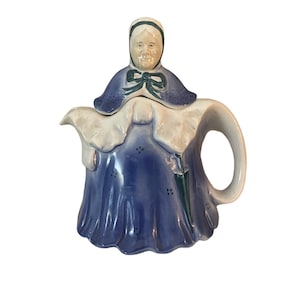 May include: A vintage ceramic teapot shaped like an elderly woman. The teapot features a blue dress, a white apron, and a bonnet. The handle and spout are white, and the face is detailed with painted features.
