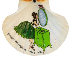 May include: A decorative seashell with a painted illustration. The artwork depicts a woman applying makeup in front of a mirror, with a green cabinet. The text "Beware the cougar in zebra's clothing" is written on the shell.