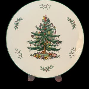 May include: A white ceramic Christmas plate with a green rim and a green and red Christmas tree design. The tree is decorated with ornaments, presents, and a bird. The plate is on a wooden stand.