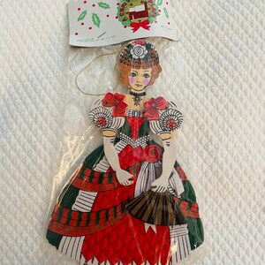 May include: A vintage Christmas ornament in the shape of a woman in a long dress, holding a fan. The ornament is made of cardboard and features a red, green, and white color scheme. The woman has red hair and is wearing a hat. The ornament is in a plastic bag.