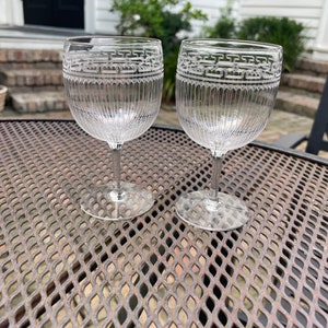 May include: Two clear glass wine glasses with a decorative etched pattern around the rim. The glasses are sitting on a metal table with a diamond pattern.