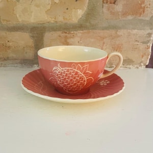 May include: A coral-coloured teacup and saucer set. The teacup features a white pineapple design and a cream-coloured interior. The saucer has a matching coral colour with a spiral design. The set is made of ceramic.