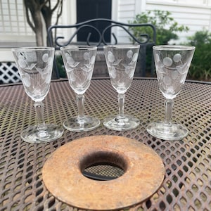 May include: Four clear glass shot glasses with etched floral designs are placed on a brown metal table with a circular metal coaster in the centre.