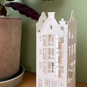 Op de afbeelding: Een gedetailleerd, wit papieren model van een gebouw met meerdere ramen, deuren en architectonische details. Het papieren model staat op een houten oppervlak, met een plant in een pot op de achtergrond. Het gebouw heeft een klassiek Europees ontwerp.