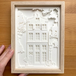 Può includere: Un'opera d'arte in carta ritagliata incorniciata raffigurante un edificio bianco dettagliato con più finestre, una porta e una bicicletta. L'opera d'arte è ambientata su uno sfondo bianco e incorniciata in una cornice di legno chiaro. La firma dell'artista e l'anno 2021 sono visibili.