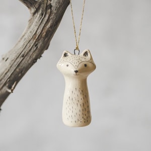 Peut inclure: Une décoration de Noël en céramique blanche en forme de renard avec des yeux noirs et un nez noir. La décoration est suspendue à une ficelle dorée sur une branche.