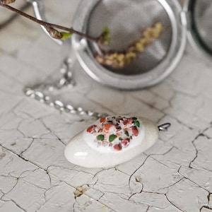 Op de afbeelding: Een roestvrijstalen thee-ei met een scharnierend deksel en ketting, naast een witte keramische hanger met kleurrijke, gespikkelde details. Een klein takje met knoppen rust in de infuser.