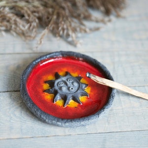 Puede incluir: Un quemador de incienso de cerámica con un diseño de sol rojo y amarillo. El quemador tiene un borde negro y un pequeño palo de incienso descansa sobre el borde.