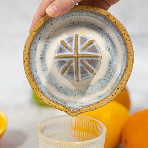 May include: A ceramic citrus juicer with a blue and white design and a brown rim. The juicer is being held over a clear glass with a ribbed pattern. There are oranges in the background.