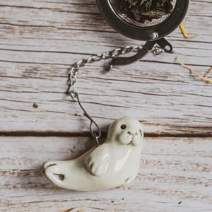 May include: A stainless steel tea infuser with a chain and a ceramic seal figurine. The infuser is filled with loose leaf tea. The seal is white with black eyes and a small nose. The background is a light brown wooden surface.