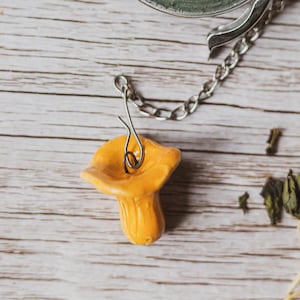 Pode incluir: Vista superior de um infusor de chá com um pequeno pingente de cogumelo laranja. O infusor é feito de metal prateado e contém folhas de chá verde secas. O pingente de cogumelo é preso por uma corrente e arame prateados. O fundo é uma superfície de madeira clara.
