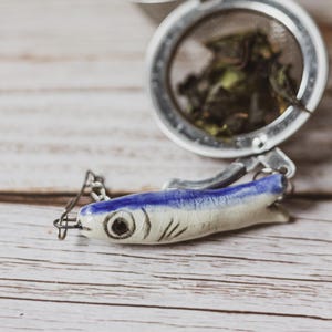 May include: A tea infuser with a ceramic fish-shaped handle, white with blue stripes and black details. The infuser is stainless steel and contains green tea leaves. It rests on a light wooden surface.