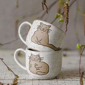 May include: Two stacked white ceramic mugs with a speckled finish. Each mug features a smiling cartoon cat illustration in tan. The mugs have a curved handle and are set against a blurred background with branches.